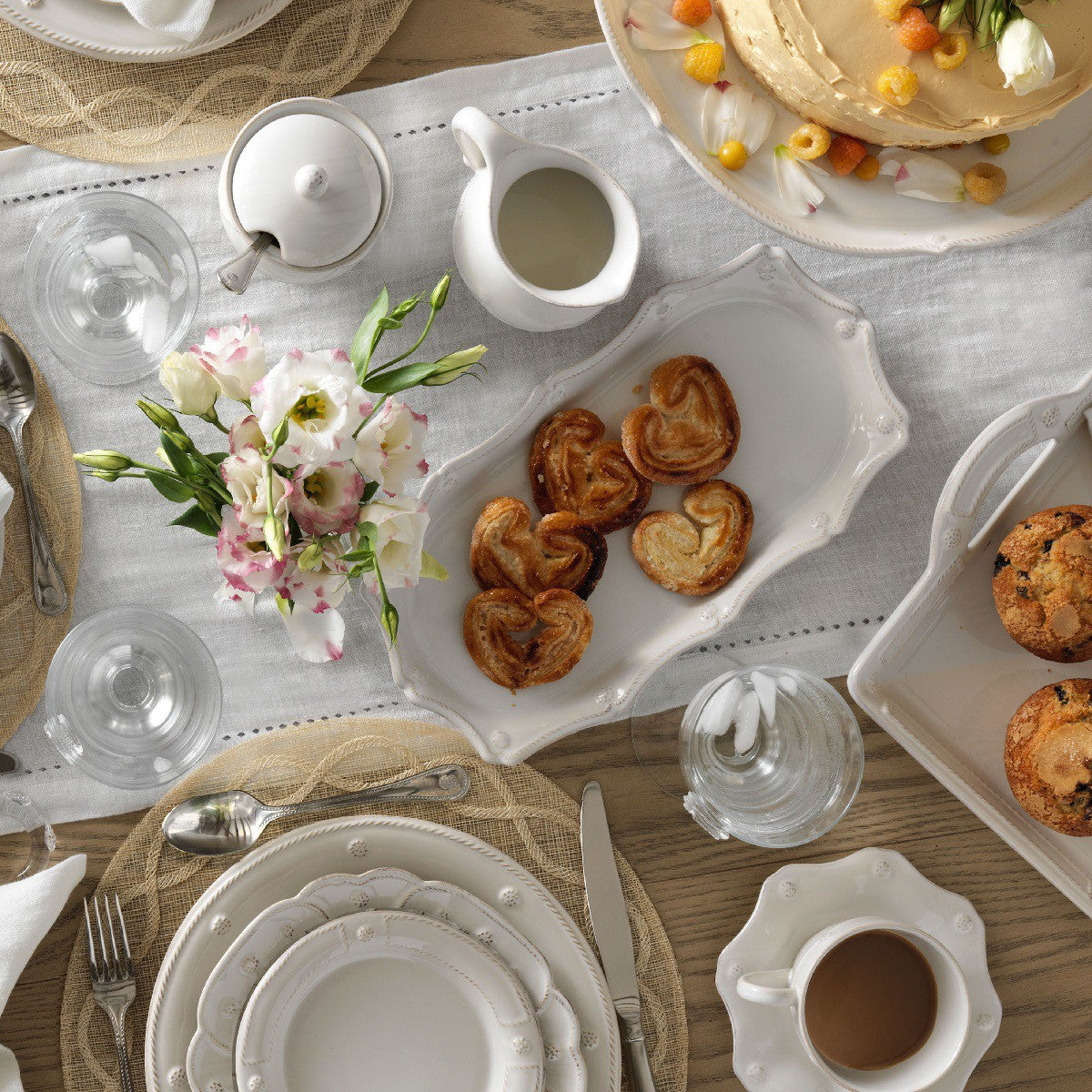 table setting with plates, glasses, coffee cup, cake stand on a wood table with a white table runner