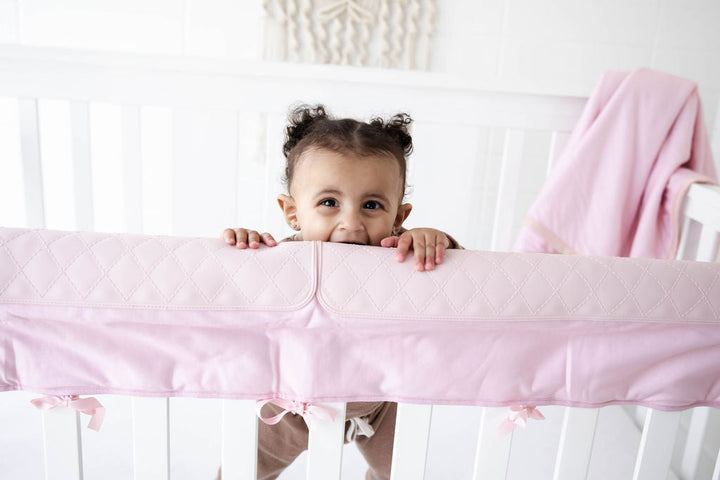 a baby girl standing and chewing on the pink crib chomper in a child's room