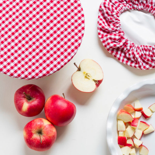 large gingham bowl cover displayed on a table next to the small cover, red apples, and pie dish with cubed apples on a white surface