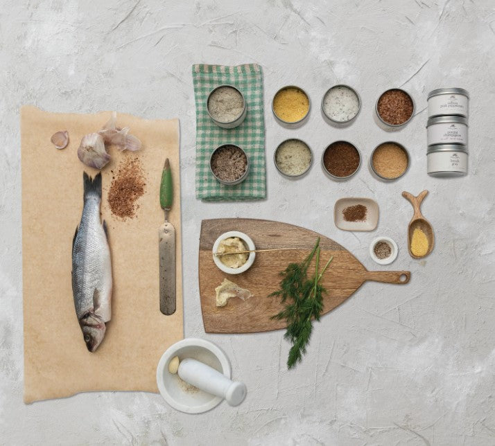 multiple open tins of seasoning displayed next to a cutting board, fish, mortar and pestle, and towel on a light rough surface