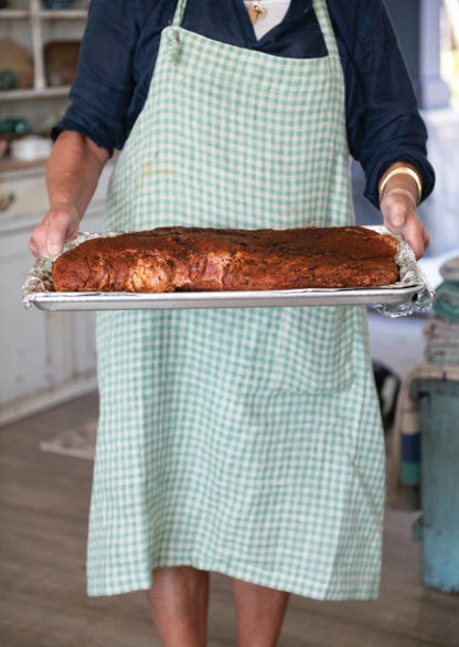a woman wearing a blue and white checked apron and holding a cooking sheet filled with seasoned meat 