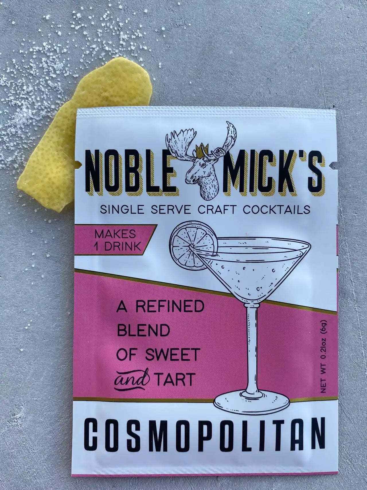 noble mick's single serve packet of cosmopolitan mix on a bar top with sugar and a lemon peel.