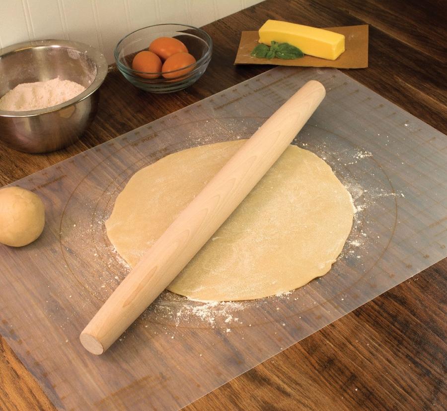 the french rolling pin displayed on a rolled out pastry next to flour eggs and butter on a dark stained wood surface