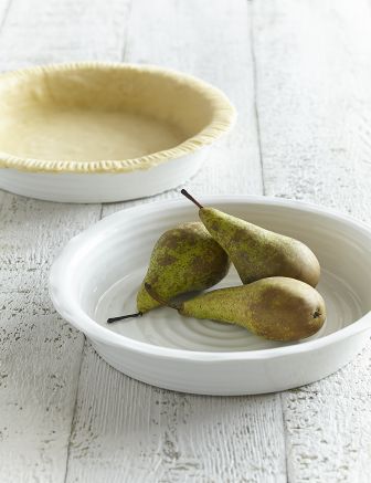 pie dish in foreground with pears in it and pie dish on background lined with pie dough, on wooden table.
