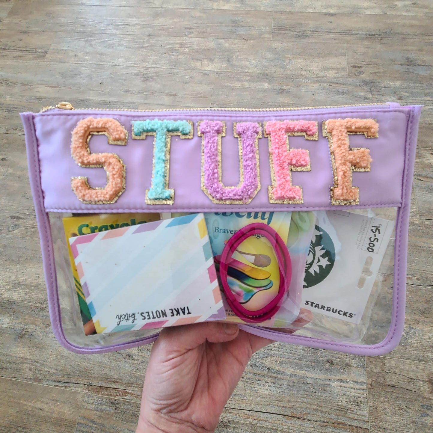 hand holding clear nylon bag with purple trim, chenille patches that spell "stuff", and filled with stuff.