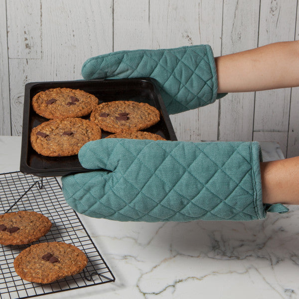 a person wearing two of the lagoon heirloom stonewash oven mitts while holding a hot pan of cookies over a white a gray marble countertops