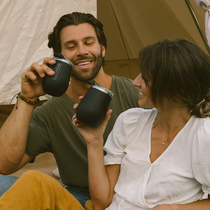 uncorked wine tumblers being held by a man and woman while sitting in front of a tent