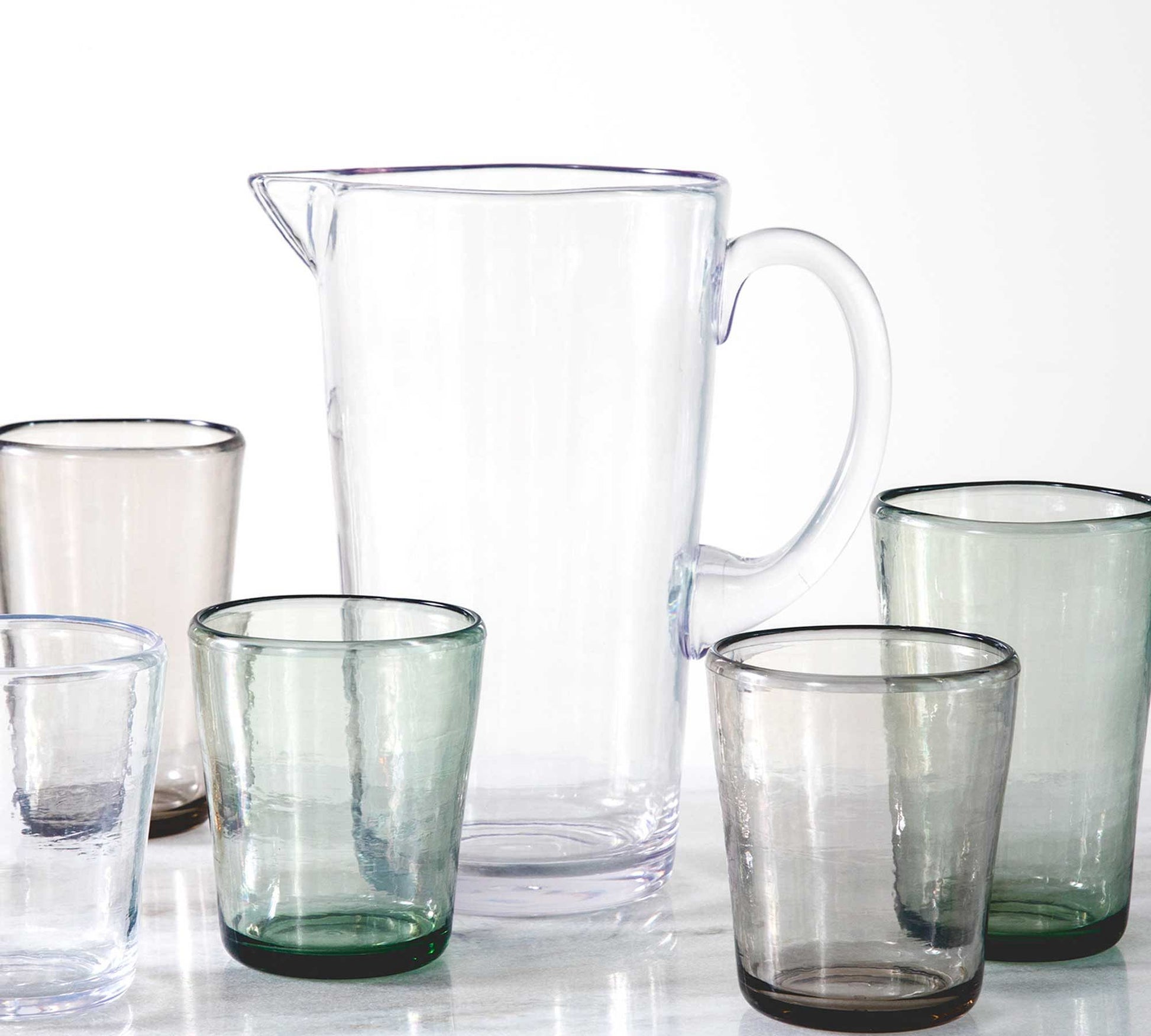 clear pitcher surrounded by assorted glasses on white background.