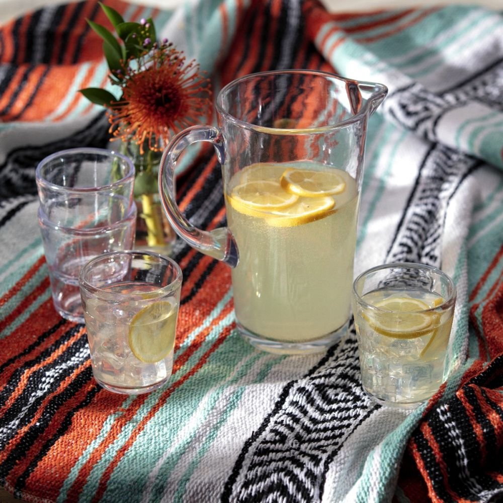 clear pitcher and glasses filled with leamonade set on a blanket with flowers.