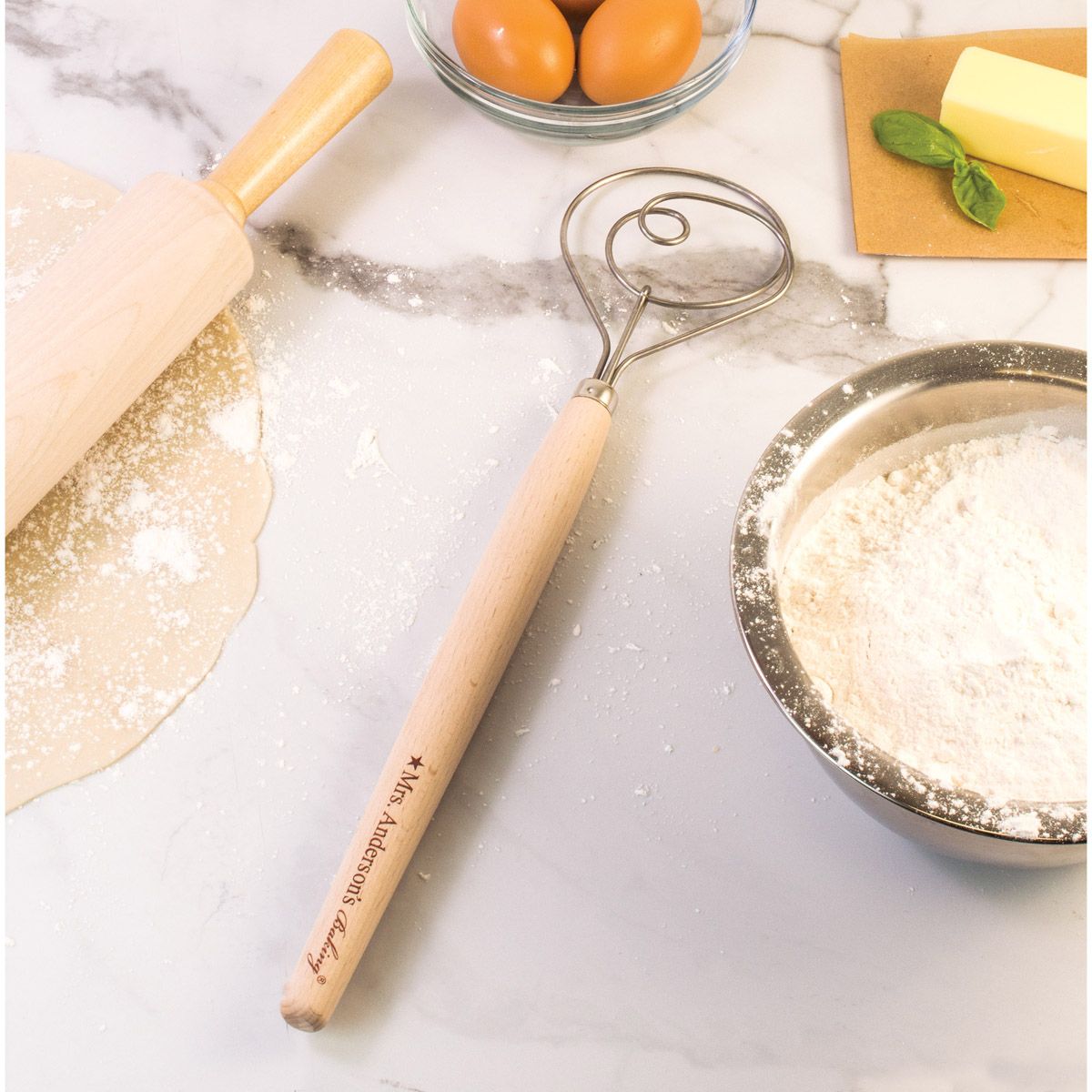 wooden handled dough whisk displayed on a white and gray marble surface next to a rolled out pastry, bowl of flower, bowl of eggs, stick of butter, and rolling pin