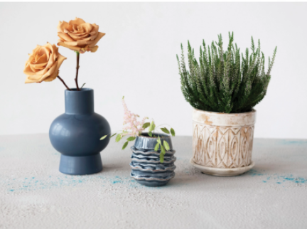 stoneware ruffled planter displayed next to a cream planter and tall blue vase with flowers inside on a white rough surface against a white background