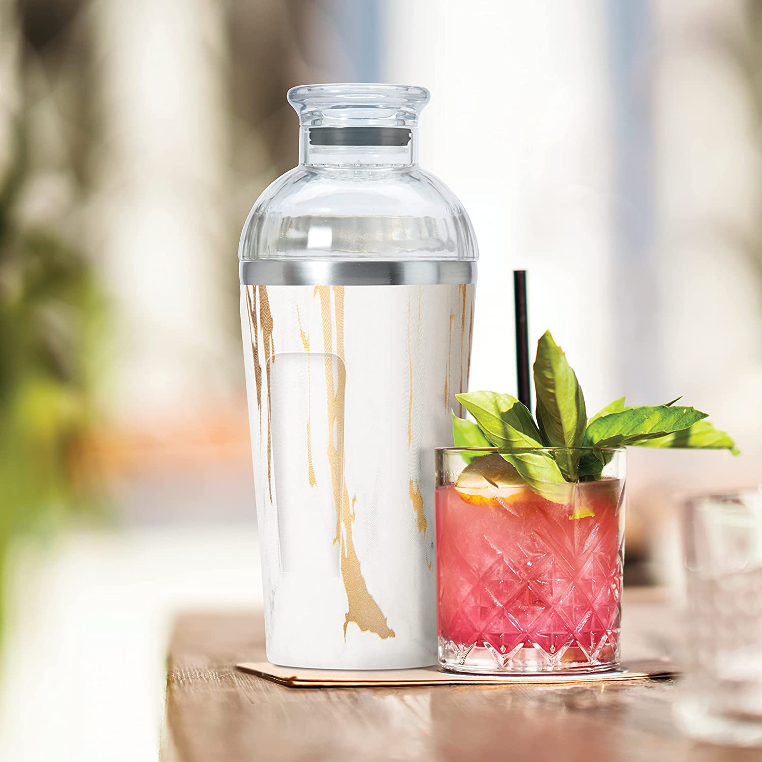 cocktail shaker set on a table with a pink cocktail that has a leafy garnish.