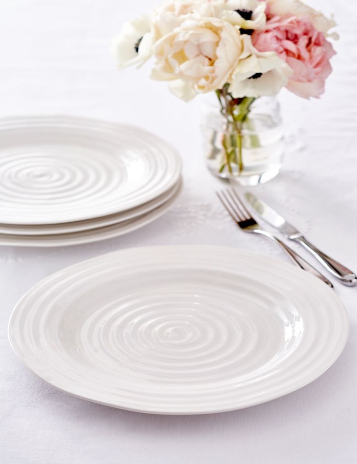 dinner plate on white table with knife and fork and stack of plates and vase of flowers in background.
