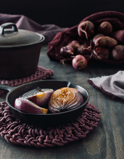 ash plum knotted trivet displayed with cast iron skillet filled with cooked onion next to a towel and cast iron dutch oven