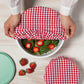 a womans hands putting the large gingham bowl cover over a bowl of strawberry salad and displayed on a whitewashed wood slat surface
